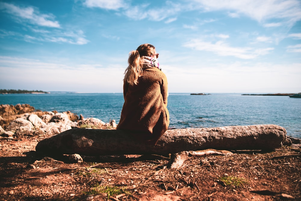 Frau im braunen Pullover sitzt tagsüber auf Felsen in der Nähe des Meeres
