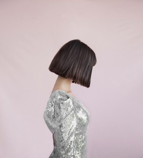 A person with a sleek bob hairstyle is wearing a shimmering silver sequin dress. The background is a soft, pale pink, creating a contrasting yet soothing effect.