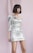 A person with a bob haircut wearing a shiny silver one-shoulder mini dress stands against a soft pink background. The dress features sequins that catch the light, giving it a reflective quality.