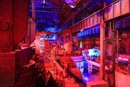 A large industrial facility filled with machinery and metal structures. The space is illuminated by a combination of red and blue lighting, creating a vibrant and futuristic atmosphere. The ceiling is high and expansive, supported by metal beams. Various industrial components and equipment are visible throughout the space, contributing to a complex and busy scene.
