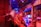 A large industrial facility filled with machinery and metal structures. The space is illuminated by a combination of red and blue lighting, creating a vibrant and futuristic atmosphere. The ceiling is high and expansive, supported by metal beams. Various industrial components and equipment are visible throughout the space, contributing to a complex and busy scene.