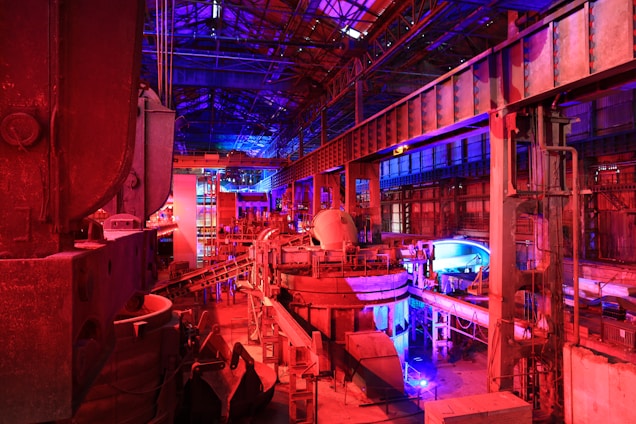 A large industrial facility filled with machinery and metal structures. The space is illuminated by a combination of red and blue lighting, creating a vibrant and futuristic atmosphere. The ceiling is high and expansive, supported by metal beams. Various industrial components and equipment are visible throughout the space, contributing to a complex and busy scene.