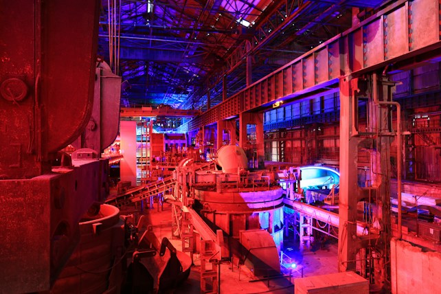 A large industrial facility filled with machinery and metal structures. The space is illuminated by a combination of red and blue lighting, creating a vibrant and futuristic atmosphere. The ceiling is high and expansive, supported by metal beams. Various industrial components and equipment are visible throughout the space, contributing to a complex and busy scene.