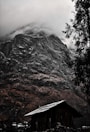 A rustic wooden cabin sits at the base of a towering, rugged mountain. The peak is shrouded in dense mist, adding a sense of mystery. Pine trees frame the scene, lending a natural touch to the rugged terrain.