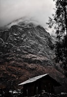 A rustic wooden cabin sits at the base of a towering, rugged mountain. The peak is shrouded in dense mist, adding a sense of mystery. Pine trees frame the scene, lending a natural touch to the rugged terrain.