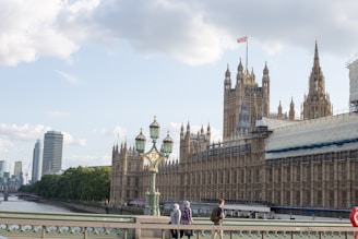 The image captures a view of the Houses of Parliament in London, featuring the intricate Gothic architecture of the building. A few people are walking across Westminster Bridge in the foreground, with a decorative lamp post visible. The sky is partly cloudy and the Union Jack is flying atop one of the towers. The River Thames flows alongside with modern buildings in the distance.