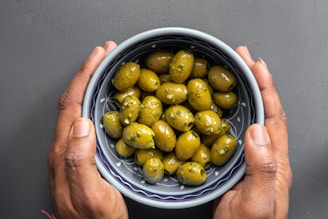 A pair of hands gently holds a bowl filled with green olives seasoned with herbs. The bowl features a blue and white pattern and is set against a dark gray background.