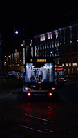 A tram illuminated by artificial lights travels through a city street at night. The tram displays the destination 'Imanta' and is surrounded by buildings with lit windows and street lamps casting a warm glow. The scene captures a quiet, urban nighttime atmosphere.