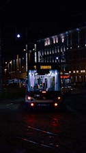A tram illuminated by artificial lights travels through a city street at night. The tram displays the destination 'Imanta' and is surrounded by buildings with lit windows and street lamps casting a warm glow. The scene captures a quiet, urban nighttime atmosphere.