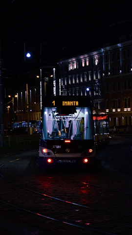 A tram illuminated by artificial lights travels through a city street at night. The tram displays the destination 'Imanta' and is surrounded by buildings with lit windows and street lamps casting a warm glow. The scene captures a quiet, urban nighttime atmosphere.