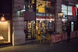 A city street scene at night, featuring the exterior of a Pizza Hut restaurant with large windows showing the interior with customers seated at tables. Bright signage is visible, and yellow outdoor tables and chairs are set up in the foreground. A neighboring store has a sale sign in the window.