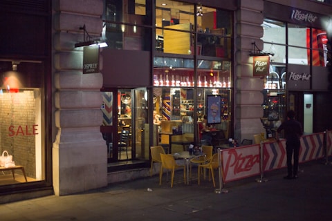 A city street scene at night, featuring the exterior of a Pizza Hut restaurant with large windows showing the interior with customers seated at tables. Bright signage is visible, and yellow outdoor tables and chairs are set up in the foreground. A neighboring store has a sale sign in the window.