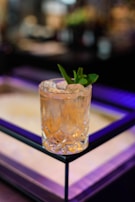 A crystal glass filled with a light amber liquid and garnished with a sprig of fresh mint sits on a reflective surface. The background features soft, blurred lights creating a bokeh effect, adding a sense of elegance and sophistication.