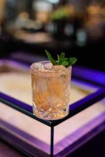 A crystal glass filled with a light amber liquid and garnished with a sprig of fresh mint sits on a reflective surface. The background features soft, blurred lights creating a bokeh effect, adding a sense of elegance and sophistication.