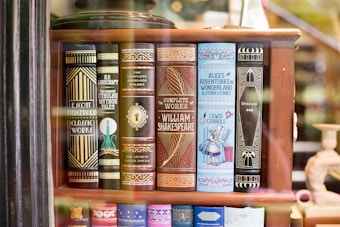A bookshelf showcasing a collection of classic literature with ornate book covers. Featured titles include works by Fitzgerald, Lovecraft, Doyle, Shakespeare, Carroll, and Stoker. The covers are elaborately designed, with some featuring illustrations like a keyhole and an image of Alice from Alice's Adventures in Wonderland.