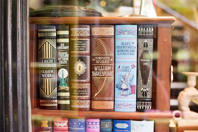 A bookshelf showcasing a collection of classic literature with ornate book covers. Featured titles include works by Fitzgerald, Lovecraft, Doyle, Shakespeare, Carroll, and Stoker. The covers are elaborately designed, with some featuring illustrations like a keyhole and an image of Alice from Alice's Adventures in Wonderland.