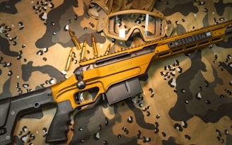 A tactical rifle with a golden-brown finish is placed on a camouflage-patterned fabric. Several bullets are arranged nearby, and a pair of protective goggles is positioned above the rifle. The overall setup appears to be militaristic, with a focus on outdoor or combat settings.