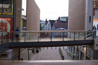 A modern outdoor shopping area with a pedestrian bridge at the center. One person stands on the bridge, using a smartphone. The setting includes glass railings, bricks, and shops with large glass windows. A mix of modern and traditional buildings can be seen in the background.