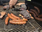 A barbecue grill is loaded with an assortment of meats, including sausages, ribs with charred edges, and a larger piece of meat that appears to have seasoning or a crust. The meats are positioned on the grates, demonstrating a well-cooked, smoky appearance.