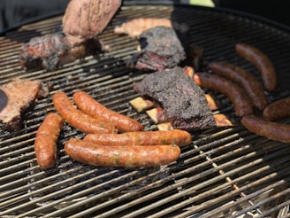 A barbecue grill is loaded with an assortment of meats, including sausages, ribs with charred edges, and a larger piece of meat that appears to have seasoning or a crust. The meats are positioned on the grates, demonstrating a well-cooked, smoky appearance.