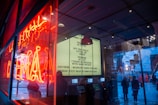 A vibrant city scene captures the reflection of a neon sign in a shop window, creating an array of glowing red hues. Inside, a theater-style marquee displays movie titles and showtimes, while the interior space features automated ticket machines. Outside, people are seen walking on a rain-slicked street, adding a dynamic urban feel.