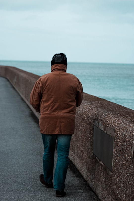 man in brown jacket and blue denim jeans standing on concrete dock during daytime in Dunkirk France