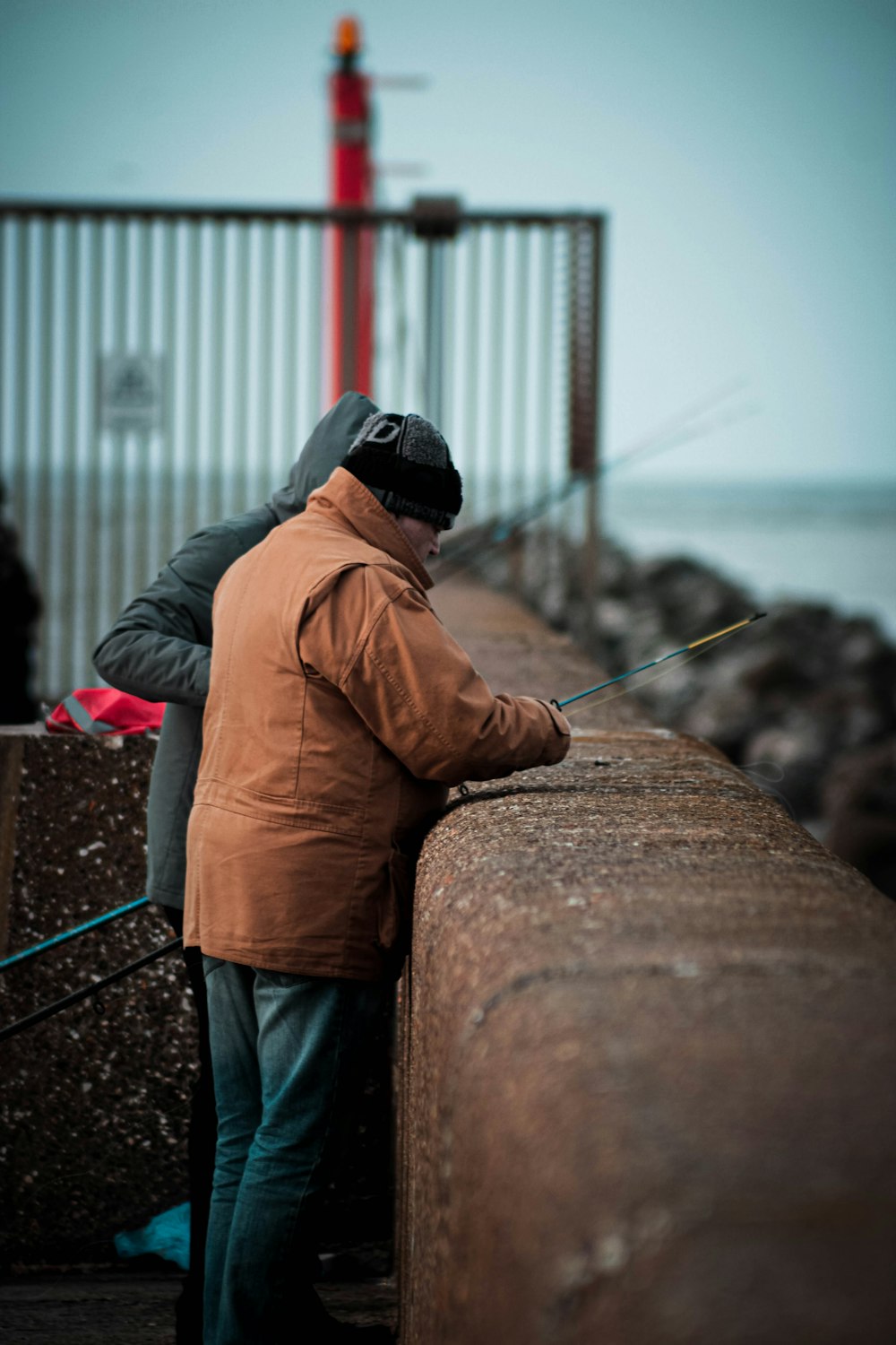 man in brown jacket and blue denim jeans standing on concrete dock during daytime