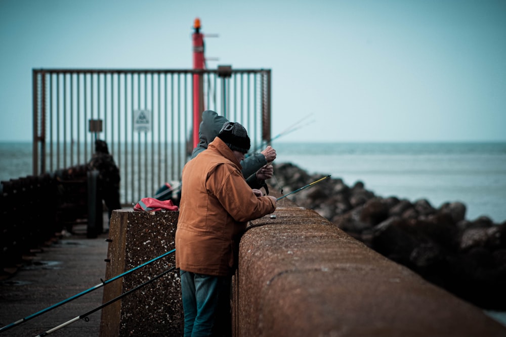 Mann in orangefarbener Jacke und schwarzer Mütze, der tagsüber auf einer Betonwand sitzt und auf das Meer blickt