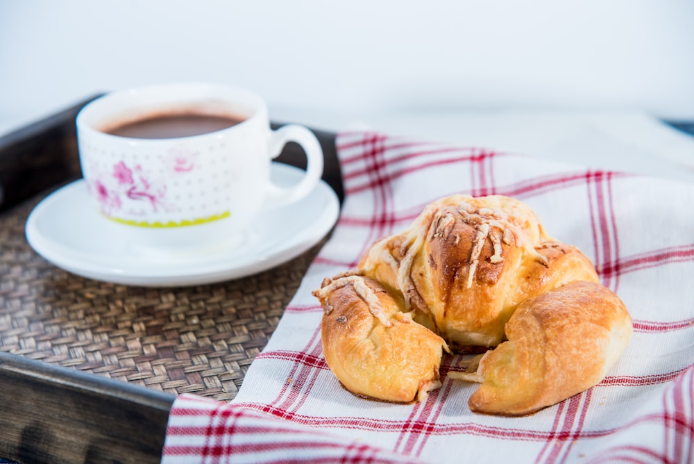 white ceramic mug with saucer beside bread on white and red checkered textile