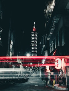 A nighttime cityscape featuring a brightly illuminated tower in the background. In the foreground, a narrow street with blurred red and white light trails creates a dynamic visual effect. Buildings line each side of the street, and there are signs with Asian script. A red lantern provides a warm glow near a building entrance.