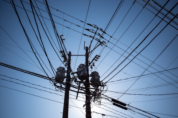A network of electrical power lines spreading out from utility poles against a clear blue sky. The poles are equipped with transformers and other electrical hardware, creating an intricate pattern of wires overhead.