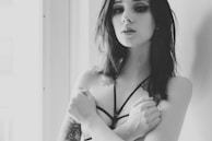 A black-and-white portrait of a person with long hair and dark eyeliner, wearing a strappy top. The person's arms are crossed over their chest, and tattoos are visible on their arm. Soft lighting comes from a window in the background, creating a gentle contrast.