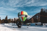 A colorful hot air balloon with the word 'Südtirol' is tethered on snowy ground. The backdrop consists of snow-covered mountains and a clear blue sky with some wispy clouds. Tall evergreen trees surround the area, giving a serene alpine atmosphere.