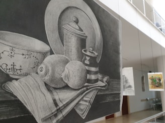 A pencil sketch depicting a still life arrangement of a bowl, a lidded jar, a striped cup, and a lemon all set on a cloth. The background is a dark shading, enhancing the contrast of the objects. The setting is in an art gallery, as indicated by other artworks hanging in the background.