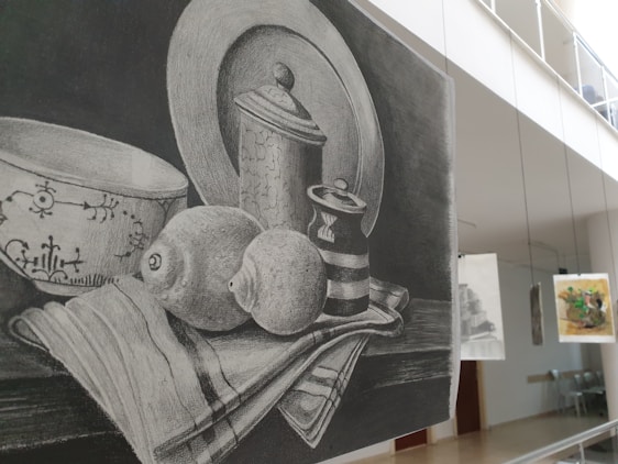 A pencil sketch depicting a still life arrangement of a bowl, a lidded jar, a striped cup, and a lemon all set on a cloth. The background is a dark shading, enhancing the contrast of the objects. The setting is in an art gallery, as indicated by other artworks hanging in the background.