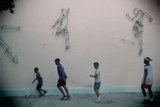 Four children are playing soccer on a concrete surface in front of a wall decorated with outline drawings of soccer players in action. The children appear to be engaged in the game, with one of them dribbling a ball. The wall provides a backdrop that enhances the dynamic feel of the scene.