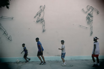 Four children are playing soccer on a concrete surface in front of a wall decorated with outline drawings of soccer players in action. The children appear to be engaged in the game, with one of them dribbling a ball. The wall provides a backdrop that enhances the dynamic feel of the scene.