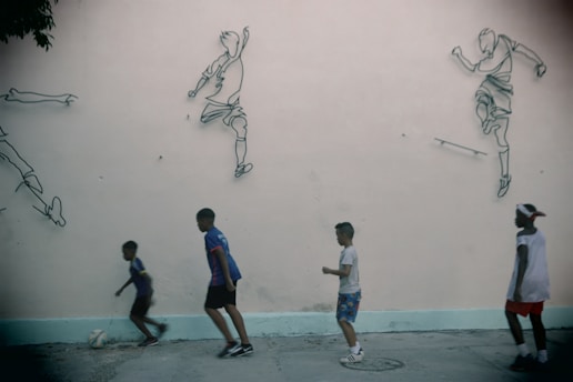 Four children are playing soccer on a concrete surface in front of a wall decorated with outline drawings of soccer players in action. The children appear to be engaged in the game, with one of them dribbling a ball. The wall provides a backdrop that enhances the dynamic feel of the scene.