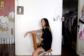 A woman with tattoos is seated on a chair in a room with a white wall. To the left, there is a decorative curtain with a sumo wrestler design. Behind her, a hanger displays clothes in an open closet. A few photos hang near the wall, while folded blankets are visible on the right.