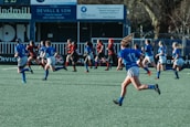 A group of athletes are running on a field, engaged in a rugby match. They are wearing blue and black sports uniforms, indicating two opposing teams. Advertisements and signs are visible in the background, suggesting a formal sports venue.