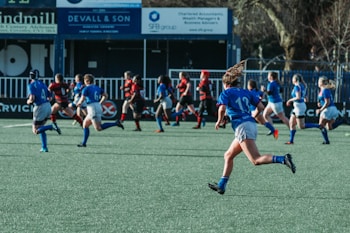 A group of athletes are running on a field, engaged in a rugby match. They are wearing blue and black sports uniforms, indicating two opposing teams. Advertisements and signs are visible in the background, suggesting a formal sports venue.