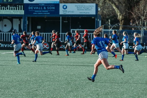 A group of athletes are running on a field, engaged in a rugby match. They are wearing blue and black sports uniforms, indicating two opposing teams. Advertisements and signs are visible in the background, suggesting a formal sports venue.