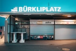 A closed kiosk with a sign reading BÜRKLIPLATZ, featuring roll-up metal shutters and advertising signs, including Lavazza branding. Two round tables stand outside, surrounded by subtle ambient lighting creating a calm evening atmosphere.