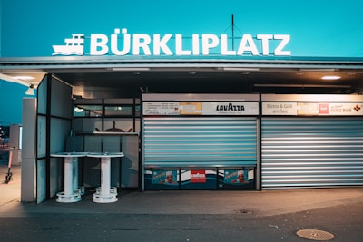 A closed kiosk with a sign reading BÜRKLIPLATZ, featuring roll-up metal shutters and advertising signs, including Lavazza branding. Two round tables stand outside, surrounded by subtle ambient lighting creating a calm evening atmosphere.