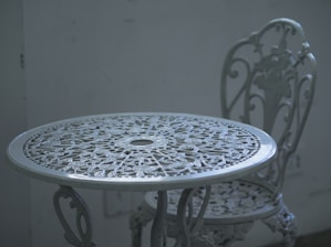 An intricately designed white metal table with a matching chair, set against a plain, muted background. The furniture features floral patterns and detailed craftsmanship, adding a vintage or classic aesthetic.