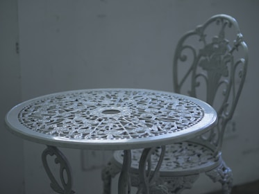 An intricately designed white metal table with a matching chair, set against a plain, muted background. The furniture features floral patterns and detailed craftsmanship, adding a vintage or classic aesthetic.