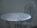 An intricately designed white metal table with a matching chair, set against a plain, muted background. The furniture features floral patterns and detailed craftsmanship, adding a vintage or classic aesthetic.