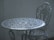 An intricately designed white metal table with a matching chair, set against a plain, muted background. The furniture features floral patterns and detailed craftsmanship, adding a vintage or classic aesthetic.
