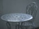 An intricately designed white metal table with a matching chair, set against a plain, muted background. The furniture features floral patterns and detailed craftsmanship, adding a vintage or classic aesthetic.