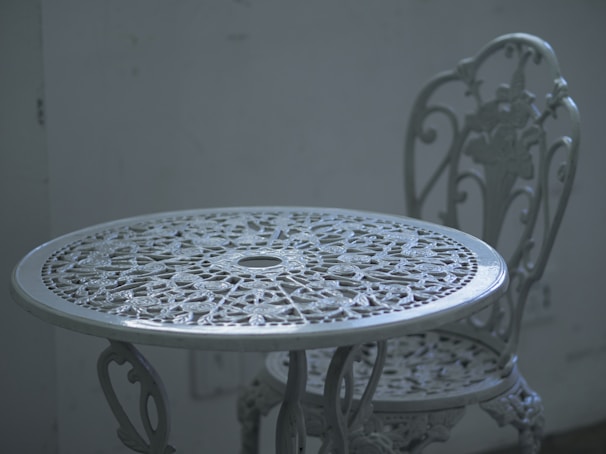 An intricately designed white metal table with a matching chair, set against a plain, muted background. The furniture features floral patterns and detailed craftsmanship, adding a vintage or classic aesthetic.
